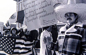Black and white photo showing people in the streets carrying both Mexican and American flags, as well as signs